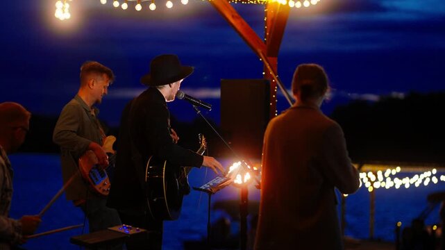 Outdoor lakeside band performing live evening concert under glowing string lights with silhouetted musicians playing instruments against twilight sky