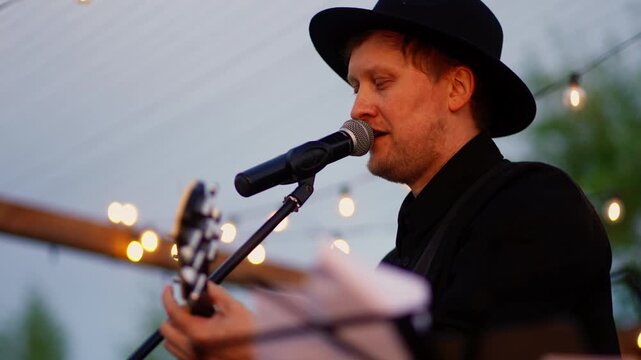 Male vocalist performing live on outdoor stage at evening event, singing into microphone under warm string lights with relaxed intimate concert atmosphere
