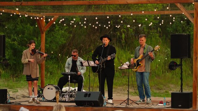 Indie band performing live on rustic outdoor stage at sunset with string lights, acoustic guitar, violin, bass and keyboard in intimate natural setting