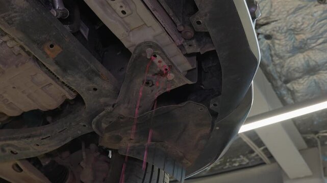 Passenger vehicle undercarriage shows red fluid dripping during automotive maintenance service. Exposed chassis and suspension components guide inspection beneath elevated vehicle in repair workshop.