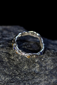 A close-up of a silver ring with a textured surface, resting on a dark stone. The ring features small gemstones embedded in its design.