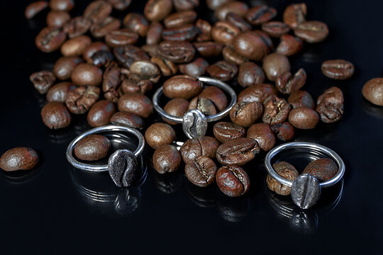 Three silver rings shaped like coffee beans on a bed of roasted coffee beans. The scene highlights a coffee theme with a dark background.