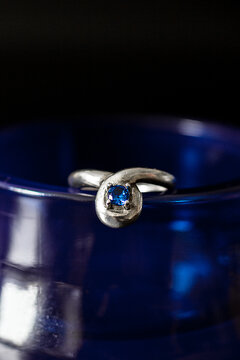 A close-up of a silver ring with a blue gemstone, placed on a dark blue glass surface. The ring features a unique twisted design.
