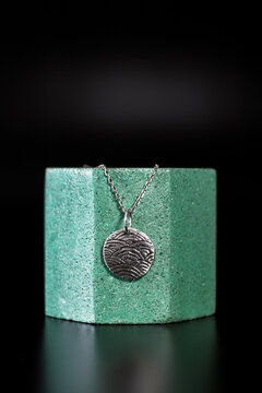 A silver fingerprint pendant hangs on a delicate chain, displayed on a textured green pedestal against a dark background.