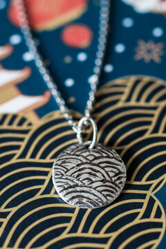 A silver pendant with a wave pattern hangs on a delicate chain. The background features a colorful, patterned surface with waves and abstract designs.