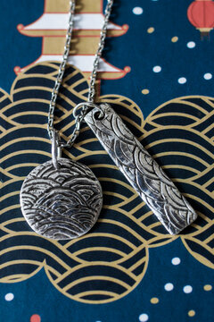 Silver jewelry featuring a round pendant and a rectangular charm with wave patterns. The background has a blue and gold wave design.