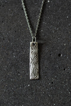 A silver pendant necklace with an intricate pattern hangs on a chain against a dark textured background. The design features a series of waves or curves.