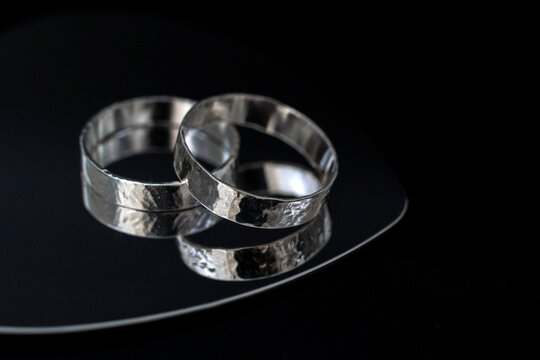 Two silver wedding rings placed on a reflective surface. The rings have a textured design and shine against a dark background.