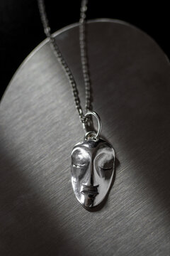A silver pendant shaped like a face hangs on a chain against a dark background. The design features a smooth, abstract representation of facial features.