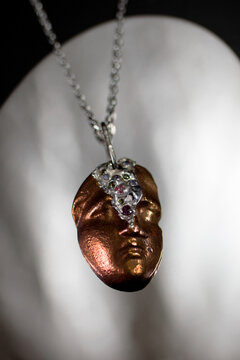 A close-up of a unique pendant featuring a bronze-colored face sculpture. The pendant is adorned with a silver chain and decorative elements.