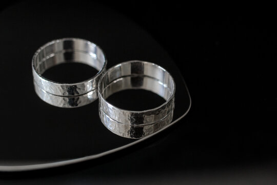 Two silver wedding bands placed on a reflective surface. The rings have a textured design and are displayed against a black background.