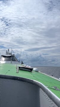View from the bow of a boat featuring a green deck, navigation equipment, and a cloudy sky above calm ocean waters, with a distant horizon line visible in the background