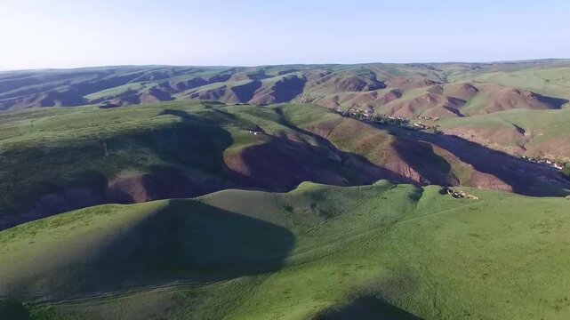Aerial drone footage of rolling green hills and rugged mountain terrain in Xinjiang, China, with scattered rural settlements and utility poles under clear blue sky