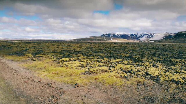 Aerial view of volcanic moss-covered plateau with distant snow-capped mountains under partly cloudy sky