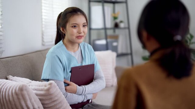 Young Asian doctor holds clipboard while listening and smiling with patient during home consultation