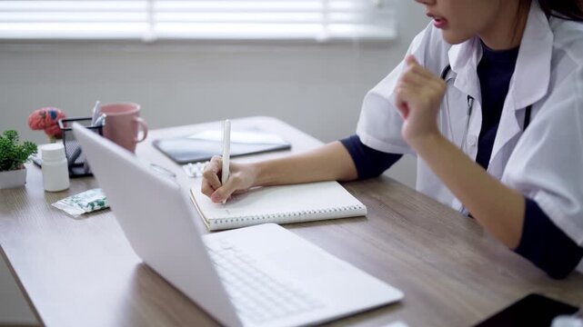 Young Asian doctor writing notes on notepad while video consulting on laptop in bright office