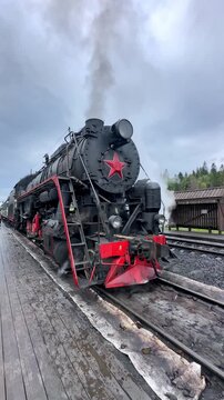 Karelia, Russia. Ruskeala Express. Vintage steam locomotive with a prominent red star emblem stands on the tracks at a railway station, releasing steam into the air. Vertical footage 