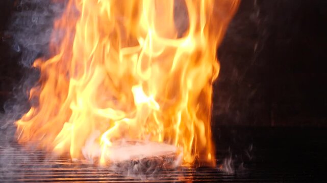 Intense orange flames and thick smoke rising over metal grill grate. Beef patty cooking on barbecue, dramatic dark background, professional restaurant kitchen process Slow motion 4k video