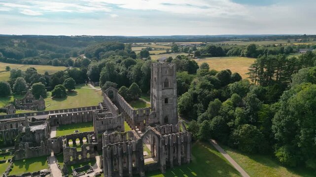 Aerial view of ancient ruins surrounded by lush greenery. Sunlight bathes stone structures, creating dramatic shadows. Fountains Abby, World Heritage Site near Harrogate North Yorkshire UK,