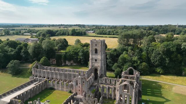 Ancient stone ruins stand tall above green fields. Tower rises proud, framing surrounding trees and grass. Fountains Abby, World Heritage Site near Harrogate North Yorkshire UK,