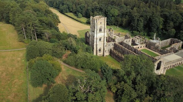 Ancient ruins stand tall amidst green fields. Trees surround the weathered stone structure. Fountains Abby, World Heritage Site near Harrogate North Yorkshire UK,
