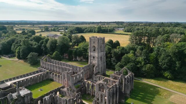 Ancient stone ruins rise above green fields and trees. Sky above is partly cloudy, casting gentle light. Fountains Abby, World Heritage Site near Harrogate North Yorkshire UK,