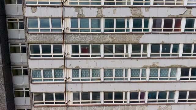 Derelict tower block in Stratford rises in an ascending drone move, revealing peeling paint, cracked plaster, stained concrete bands, and rows of aging windows on a neglected high-rise exterior.