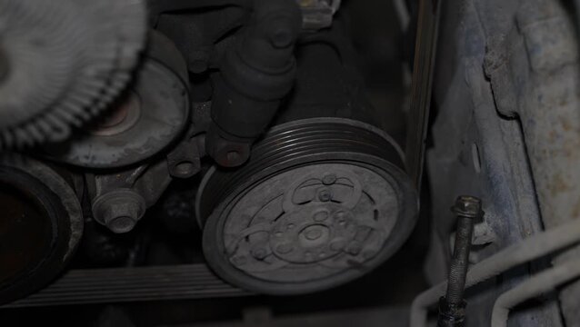 A close-up shot shows a dusty car air conditioning compressor clutch and pulley with drive belts inside the engine compartment.