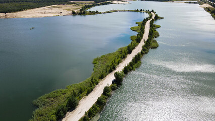 Obraz premium Industrial landscape showing a winding road separating two vast man made lakes, surrounded by green vegetation and evidence of sand quarry operations under a bright summer sky