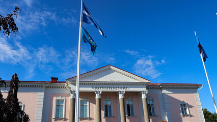 Keila Kultuurikeskus building stands under a clear blue summer sky, showcasing its architectural facade with columns and windows, as European Union and Estonian flags proudly wave in front © Dark Artistic