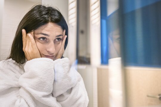 Brunette caucasian woman looks to mirror with holding hands on her face concerned look. Skin aging and care. Tbilisi.Georgia