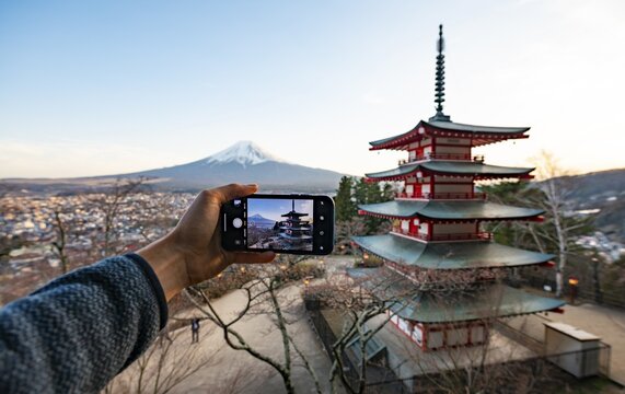 Hand holding smartphone, photographing five-story pagoda of a Shinto shrine, Chureito Pagoda, with views of Fujiyoshida City and Mount Fuji volcano at sunset, Arakura Fuji Sengen Shrine, Arakurayama Sengen Park, Yamanashi Prefecture, Japan