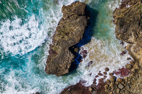 Description: Downward aerial view of ocean waves crashing onto rugged rock formations along the Yamba coastline, creating white foam and natural patterns beside the Pacific Ocean.