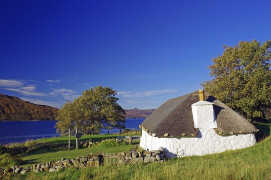 Small white house with thatched roof by the lake, surrounded by meadows and trees under a blue sky, Cottage, Holiday, Autumn, October, Hebrides, Isle of Skye, Hebrides, Scotland, United Kingdom