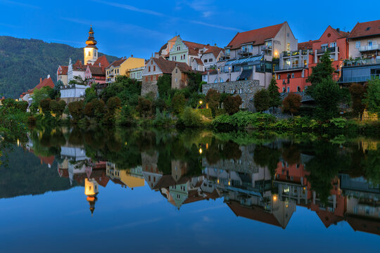 Blue hour twilight view of the illuminated historic town skyline of Frohnleiten with the parish church tower reflected in the Mur River in Styria, Austria.