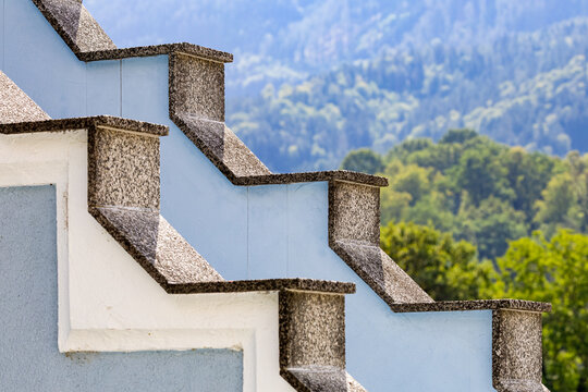 Detail of a stepped crow-step gable roofline on a light blue historic building set against forested mountain slopes in Frohnleiten, Styria, Austria