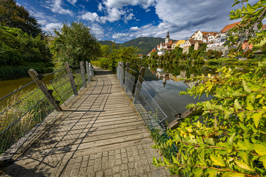 View from a wooden footbridge across the Mur River leading toward the historic town skyline and parish church tower of Frohnleiten in Styria, Austria
