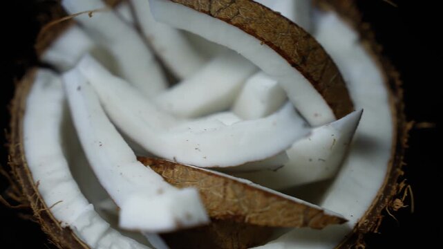 Sliced fresh coconut pieces inside the shell on a dark background, showing white flesh and brown husk texture