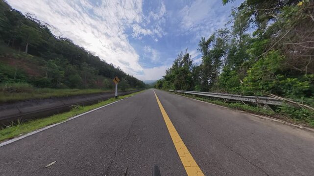 Scenic road through dense tropical forest with a yellow center line, guardrail and distant mountains beneath a dramatic blue sky.