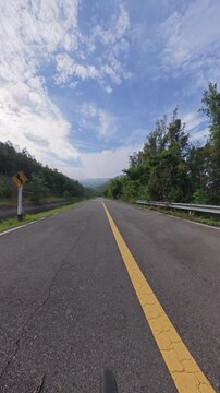 Scenic mountain road through dense forest with a yellow center line, guardrail and distant misty hills beneath a blue sky with textured clouds.