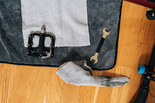 Bicycle pedal, wrench, cleaning cloth resting on dark towel on wooden floor. Concept of process of maintaining and repairing bike components, close-up, cropped shot, top view, nobody, no people.