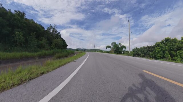 Scenic mountain road curving through lush green countryside with a visible cyclist shadow, distant hills and a blue sky filled with soft clouds.