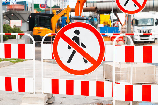 A construction site is visible with barriers and no pedestrian signs in the foreground. Heavy machinery and construction equipment are present in the background, indicating ongoing work