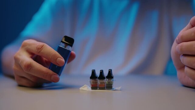 Male hands assembling electronic cigarette by inserting refillable liquid cartridge into battery device, with colorful e juice pods on table for flavorful smoking alternative, close-up, slow motion.