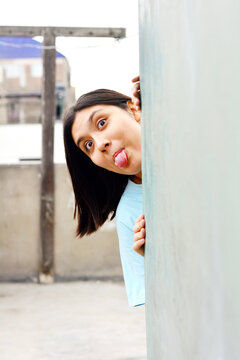 Teenager latin girl peeking behind the wall and sticking the tongue out.