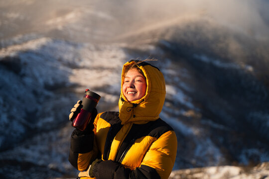Portrait of smiling woman with thermos cup in snowy mountains, enjoying sunrise light, outdoor lifestyle and travel concept.