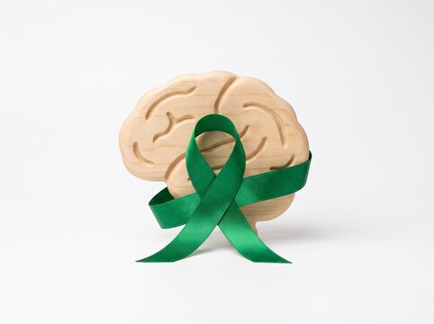 A wooden brain model with a green awareness ribbon wrapped around it, symbolizing mental health or neurological support on a white background.