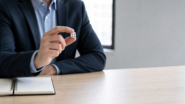 Businessman in a suit holding a pair of dice while seated at a wooden table with a notebook and window in the background, symbolizing chance and decision-making