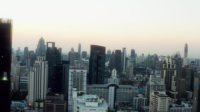 Drone pans across a massive dark skyscraper above Bangkok at early sunrise.