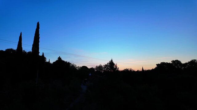Sunrise timelapse of two tall cypress trees silhouetted against a gradient sky, as the sun rises above a quiet countryside horizon